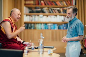 Close up shot from the side of Ringu Tulku Rinpoche seated on the stage speaking to aa male member of the audience who is stood in front of him